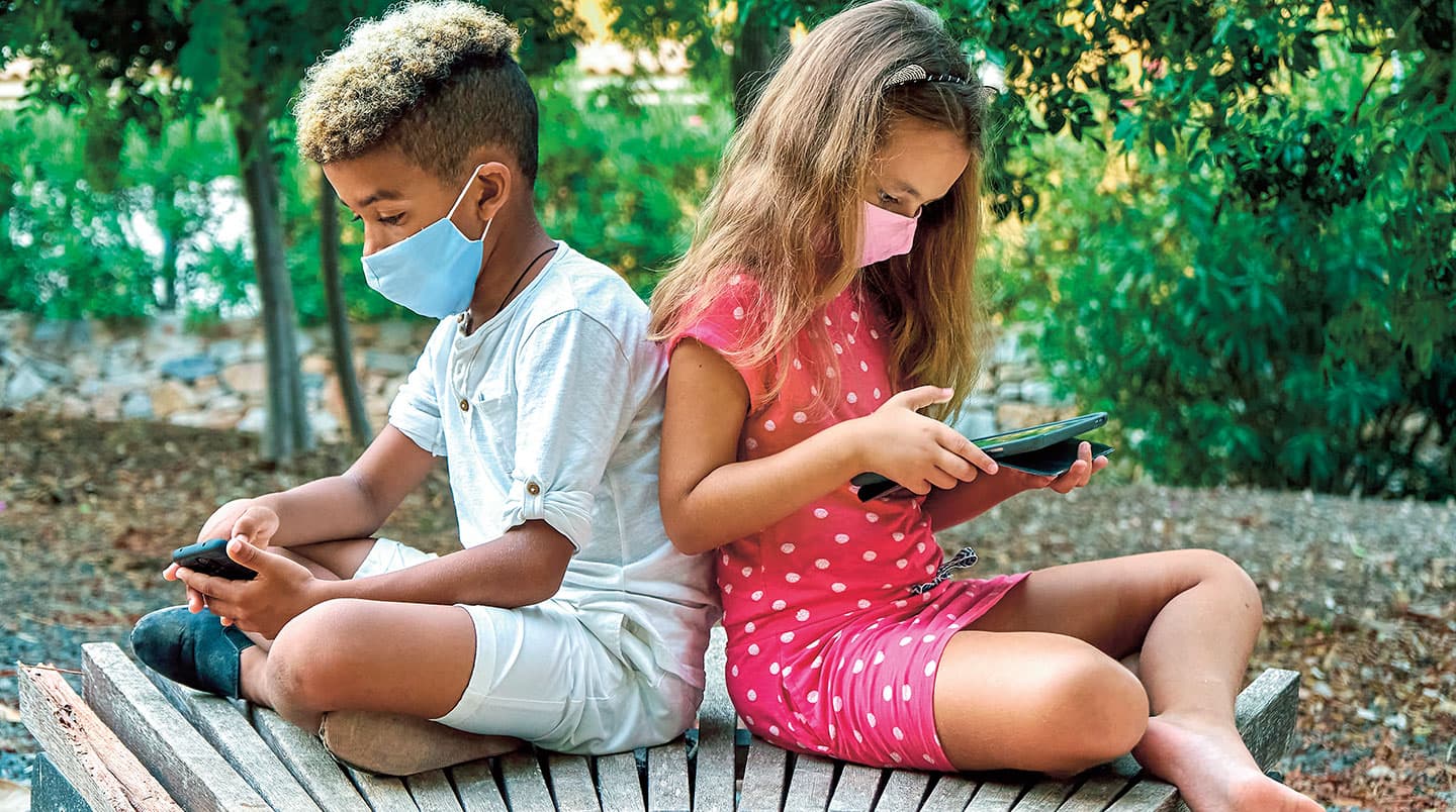 Two young kids sit with their backs to each other as they look at their tablet and phone.