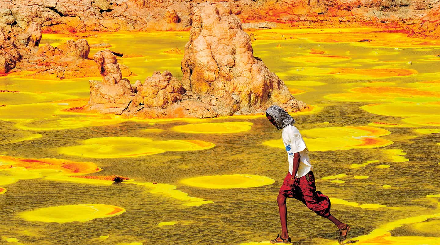 A boy walks across brightly colored sand near salt mounds and toxic puddles.