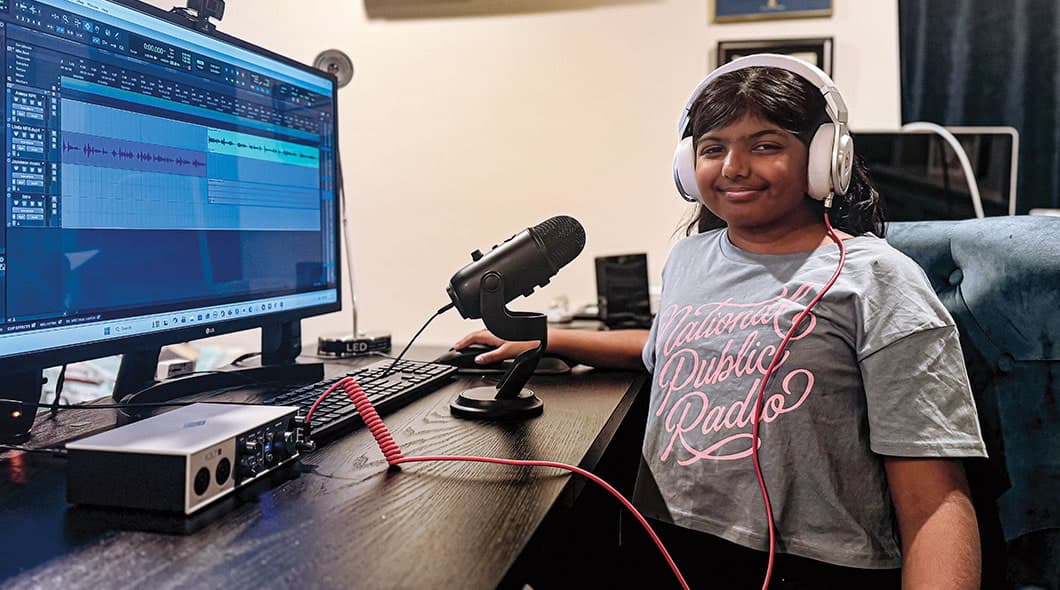 A girl wearing headphones sits at a desk with audio editing software and a microphone