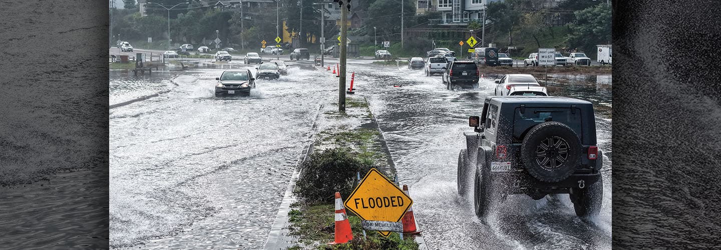 Vehicles drive through a flooded street with a yellow sign reading FLOODED in the foreground