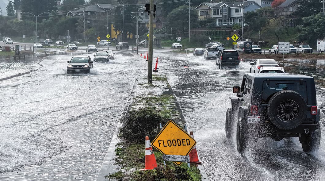 Cars drive through flooded intersection with yellow warning sign reading Flooded