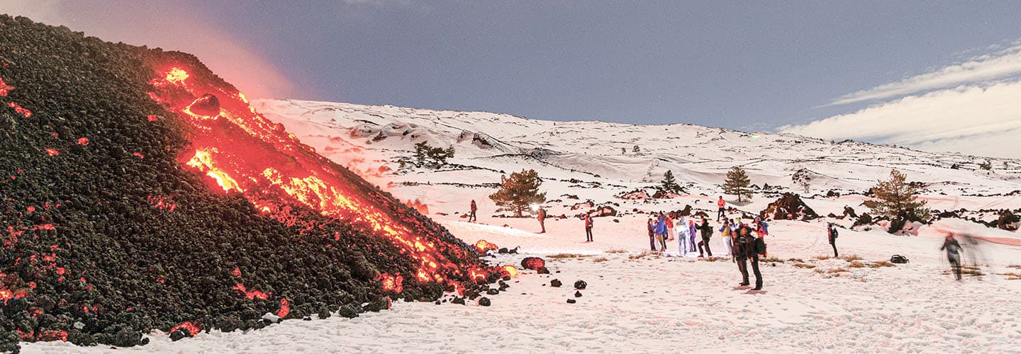 People observe glowing red lava flowing down a snow-covered volcanic slope