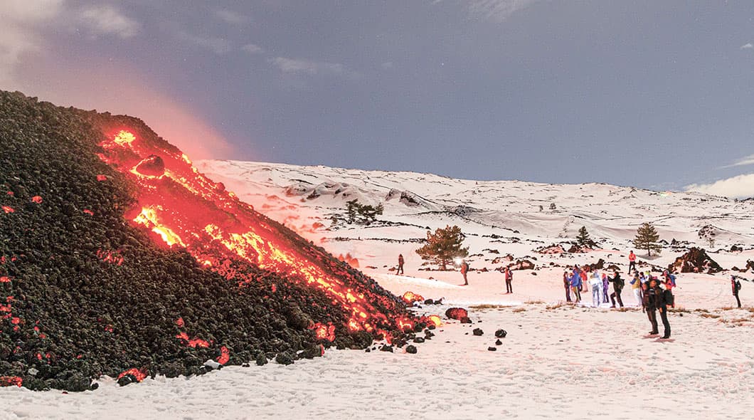 Glowing red lava flows down a dark volcanic slope as people observe from a snowy landscape