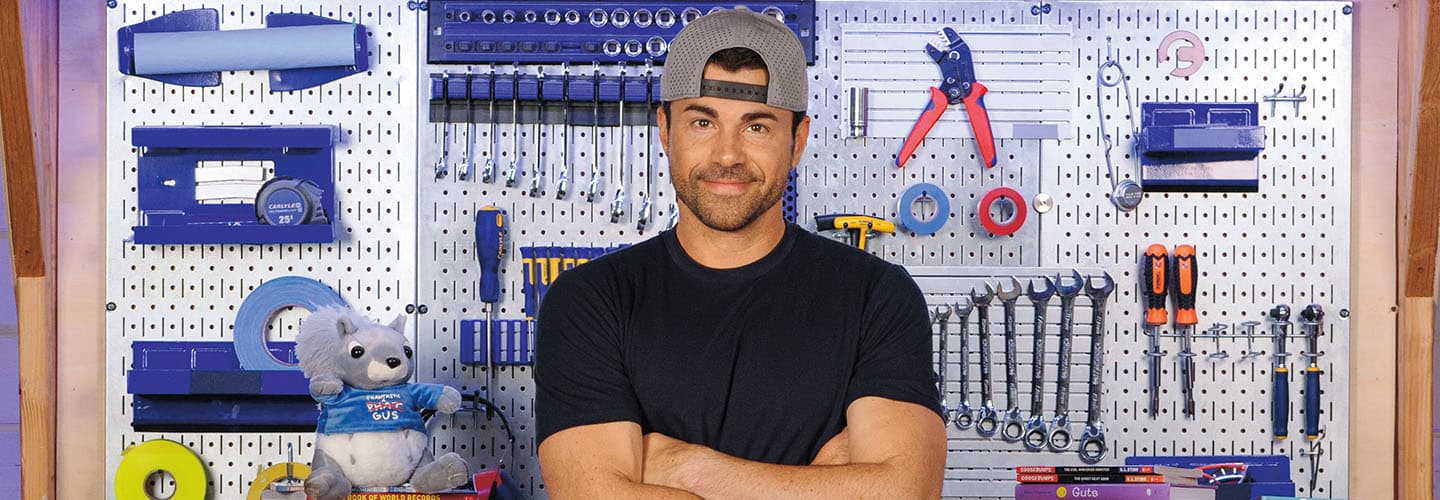 A man stands with arms crossed in front of a pegboard wall covered with organized tools