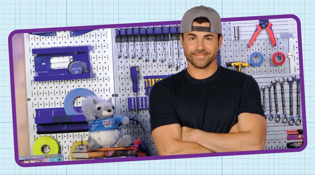 A man stands with arms crossed in front of a pegboard wall lined with tools and a stuffed animal