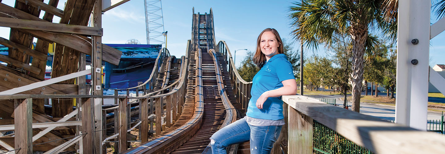 A woman leans against a railing beside a large wooden roller coaster at an amusement park