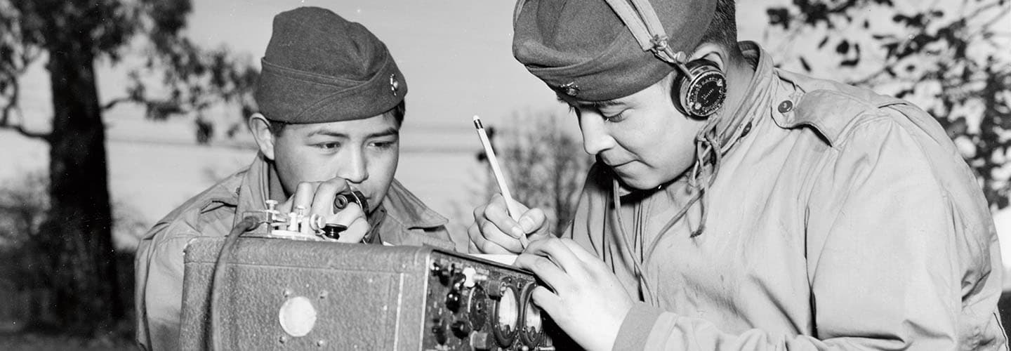 Historical photo of a soldier speaking into a radio handset as another in headphones writes notes