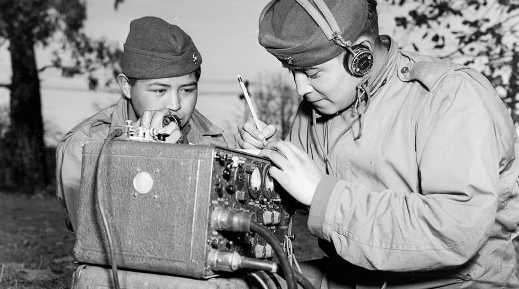 Historical photo of a soldier speaking into a radio handset as another in headphones takes notes